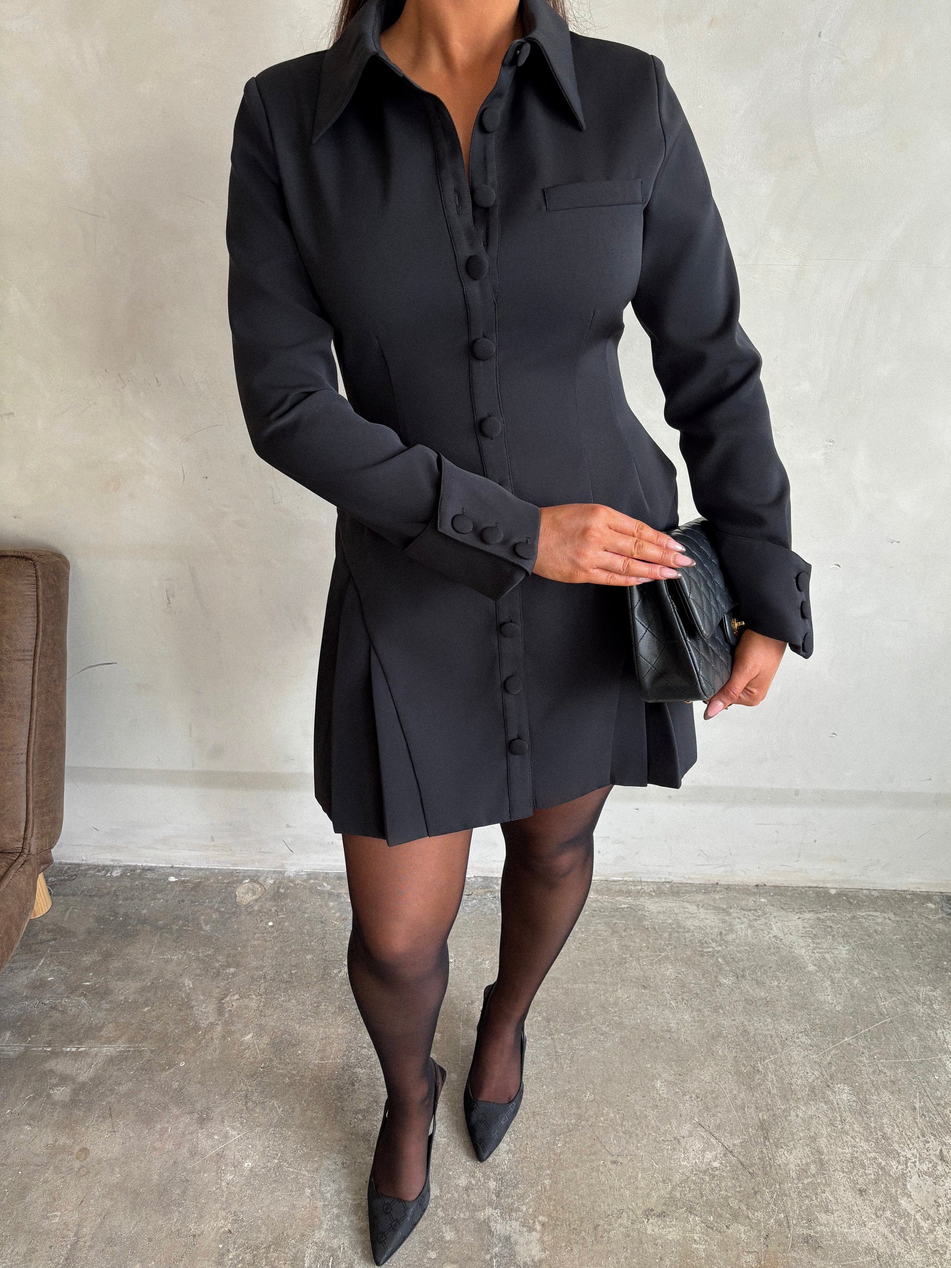 Close-up of Ardore London black structured long sleeve pleated mini dress, showcasing sculpted curved seam pleats, angled triple-buttoned cuffs, and a dramatic 1970s-inspired collar—crafted from soft, structured fabric for a timeless winter-ready silhouette with full lining and hidden bust button for flawless comfort.